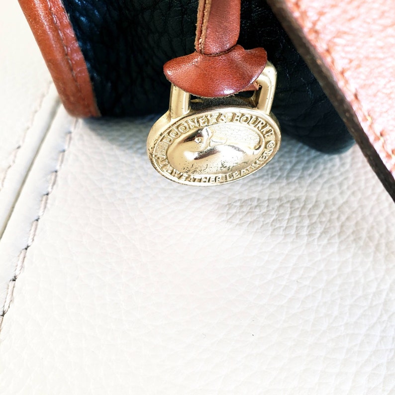 May include: Close-up of a gold-colored Dooney & Bourke brand tag with a small, embossed rabbit logo. The tag is attached to a brown leather strap.