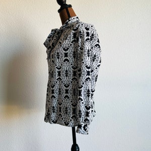 May include: A white blouse with a black and white geometric pattern. The blouse has a bow tie at the neck and long sleeves.
