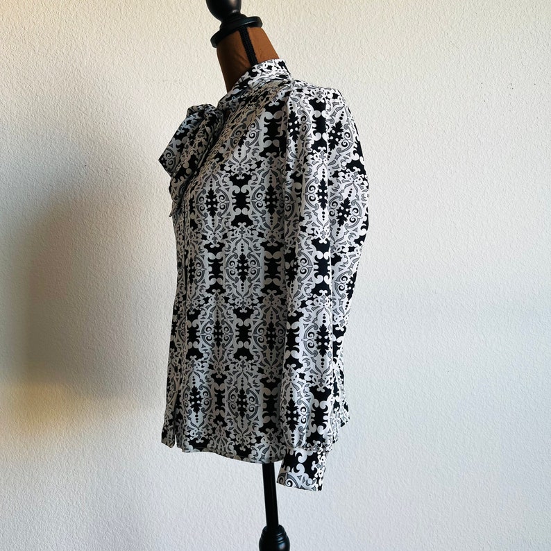 May include: A white blouse with a black and white geometric pattern. The blouse has a bow tie at the neck and long sleeves.