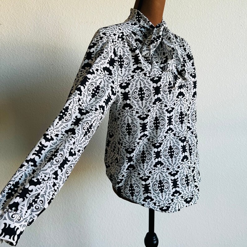 May include: A white and black patterned blouse with a high neck and a bow tie. The blouse has long sleeves and a fitted silhouette.