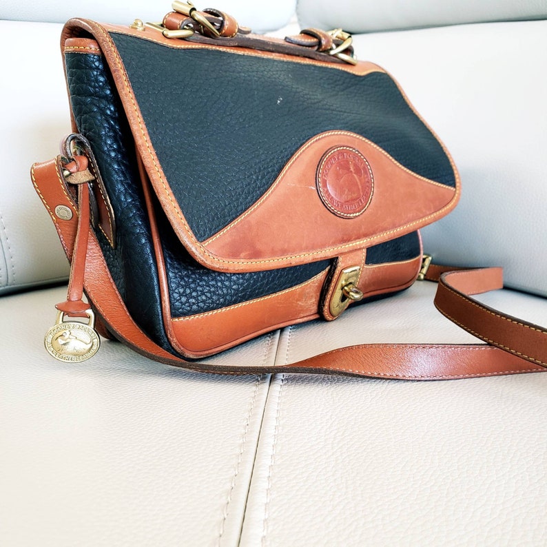 May include: A black and brown leather shoulder bag with a flap closure and a brass buckle. The bag has a long adjustable strap and a small circular charm with a logo.
