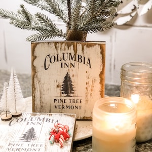 May include: A rustic wooden block planter with the text "Columbia Inn Pine Tree Vermont" holds a small artificial pine tree. A smaller wooden sign with the same text and a bundle of candy canes sits in front. Lit candles in glass jars add warmth.
