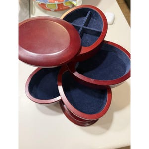 May include: A multi-tiered, oval-shaped wooden jewelry box with a dark cherry finish. The box features several rotating compartments lined with deep blue velvet, including one top section divided into four smaller quadrants for organization.