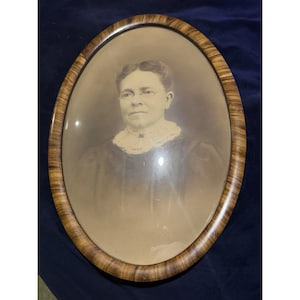 May include: An oval-shaped portrait featuring a sepia-toned photograph of a person wearing glasses and a high-collared lace garment. The portrait is encased in a convex glass cover and framed by a polished wooden border with a distinct grain pattern.