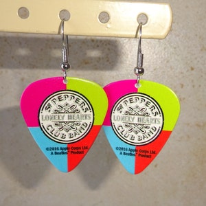 May include: A pair of guitar pick-shaped earrings. Each earring is divided into four colored sections: pink, green, blue, and red. The center features a circular design with the text "Sgt. Peppers Lonely Hearts Club Band".