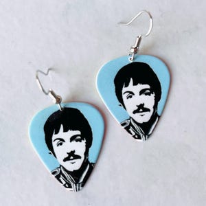 May include: A pair of light blue guitar pick earrings featuring a black and white portrait. The earrings have a silver hook and are a unique accessory for music lovers.