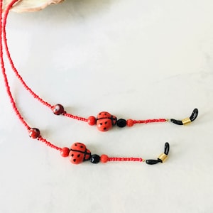 May include: Red beaded eyeglass chain with ladybug accents. The chain features small red beads, ladybug-shaped beads with black details, and black and gold-toned connectors for the glasses. A decorative and functional accessory.