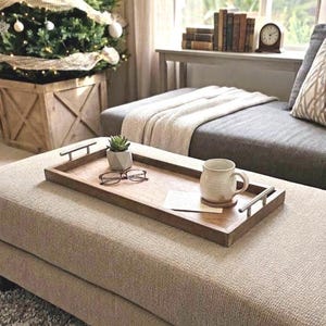 May include: A wooden serving tray with metal handles holds a small succulent in a geometric pot, eyeglasses, a mug, and a coaster. The tray sits on a beige ottoman in front of a couch and a Christmas tree.