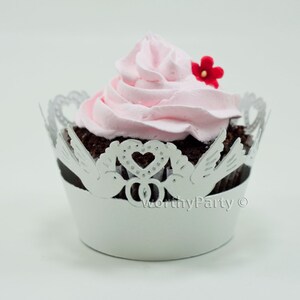 May include: White cupcake liner with a silver lace design featuring two doves and a heart with rings. The cupcake is topped with pink frosting and a red flower.