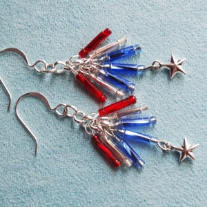 May include: A pair of silver earrings with red, white, and blue glass beads and silver star charms. The earrings are designed to look like fireworks.