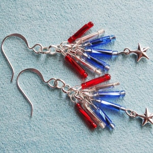 May include: A pair of silver earrings with red, white, and blue glass beads and silver star charms. The earrings are designed to look like a firework.