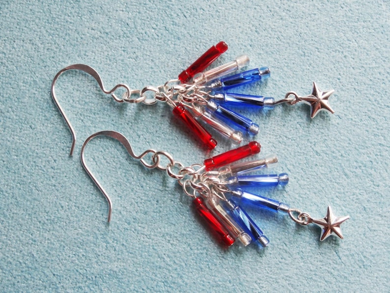 May include: A pair of silver earrings with red, white, and blue glass beads and silver star charms. The earrings are designed to look like a firework.