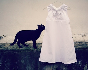 May include: A black cat looks at a white sleeveless dress with a white collar hanging on a hanger against a white wall.