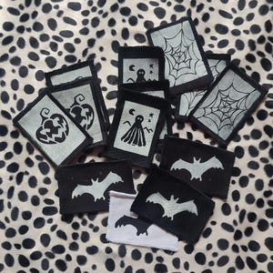May include: A collection of Halloween-themed patches featuring various designs. The patches include images of bats, spiderwebs, ghosts, and jack-o'-lanterns. The patches are black and white, with a silver-colored design. The patches are arranged on a spotted background.