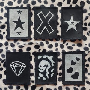 May include: Six black and white patches with various designs. The designs include a star, an X, a diamond, a skull, and hearts. The patches are rectangular with a black border and are set against a spotted background.