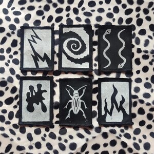 Può includere: Sei toppe nere e argentate con vari disegni: fulmine, spirale, due serpenti, macchia astratta, insetto e fiamme. Le toppe sono disposte su uno sfondo di tessuto maculato.