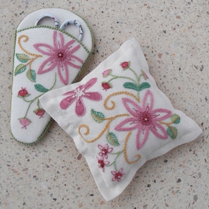 May include: A white fabric pincushion with pink and yellow embroidered flowers. A white fabric scissor holder with pink embroidered flowers and a pair of silver scissors is also included.