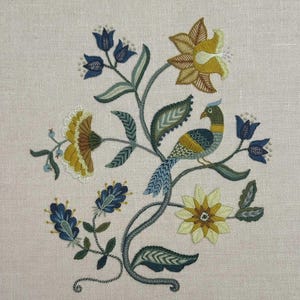 May include: An embroidery design on light beige fabric featuring a central bird with blue, yellow, and green plumage perched on a curved stem. The bird is surrounded by various stylized flowers in shades of deep blue, mustard yellow, and sage green, with intricate leaf patterns and delicate stitching throughout the composition.