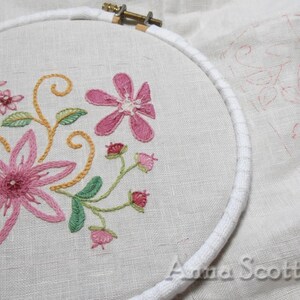 May include: A close-up of a floral embroidery design in progress, featuring pink and green flowers and leaves, stitched on a white linen fabric in a circular embroidery hoop. The design is partially completed, with some areas still outlined in pink.