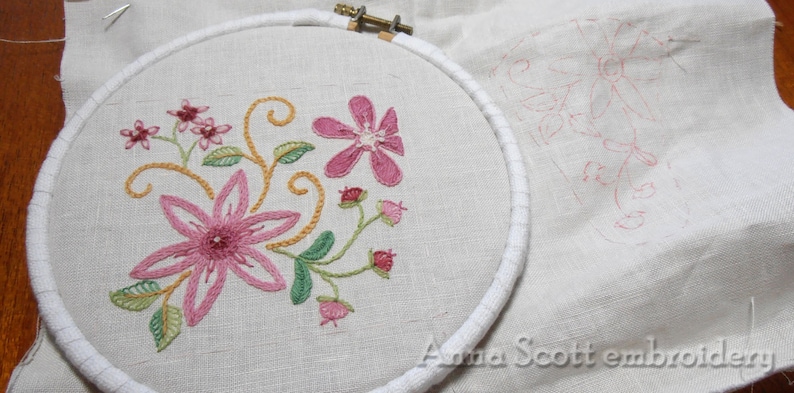 May include: A close-up of a floral embroidery design in progress, featuring pink and green flowers and leaves, stitched on a white linen fabric in a circular embroidery hoop. The design is partially completed, with some areas still outlined in pink.