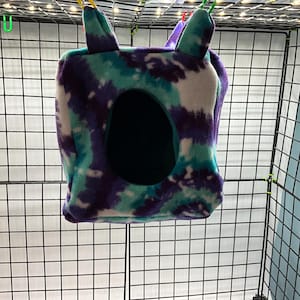 Puede incluir: Un escondite para mascotas de forro polar teñido anudado en tonos turquesa, morado y blanco. El refugio en forma de cubo tiene una gran abertura circular y dos protuberancias en forma de oreja en la parte superior. El escondite está suspendido dentro de una jaula de alambre.