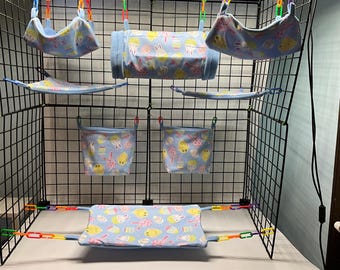 May include: A blue wire cage with multiple hanging fabric accessories for small pets. The accessories feature a light blue background with a yellow, pink, and white pattern of bunnies and cupcakes.