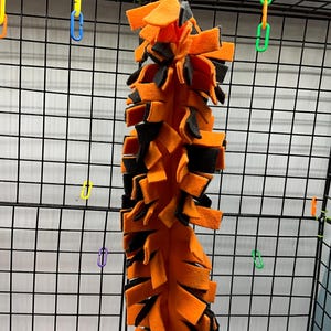 May include: Orange and black fabric toy for pets. The toy is made of strips of fabric that are tied together. It is hanging from a cage.