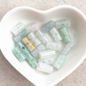 Pode incluir: Uma coleção de contas de jade verde, branco e amarelo em um prato de cerâmica branco em forma de coração. As contas são cilíndricas e têm um design talhado.