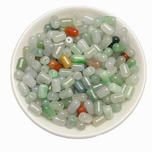 Pode incluir: Uma tigela branca cheia de contas de jade de várias cores. As contas são cilíndricas com extremidades arredondadas e incluem verde, branco, vermelho e amarelo. Algumas contas têm uma aparência translúcida.