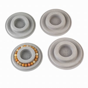 May include: Four gray, circular, tiered display stands for bracelets. The stands have a raised center circle with a measurement printed in centimeters. One stand has a bracelet of orange and yellow beads on it.