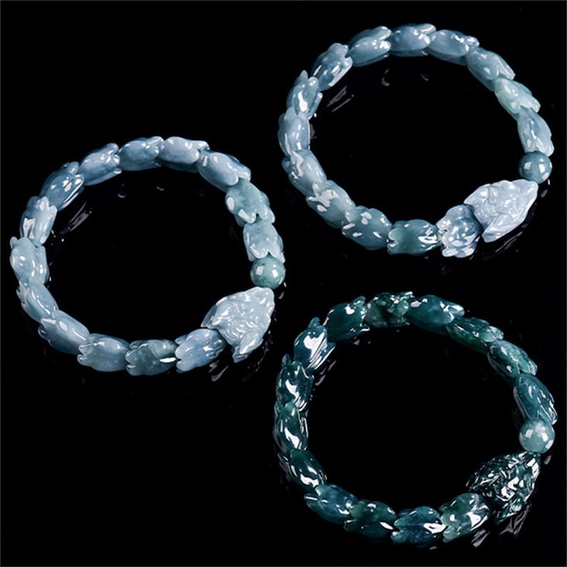 Puede incluir: Tres pulseras de jade en tonos azules y verdes se muestran sobre un fondo negro. Cada pulsera presenta un patr&oacute;n repetitivo de formas de animales talladas, con una talla de animal m&aacute;s grande como punto focal. Las pulseras son probablemente para usar en la mu&ntilde;eca.
