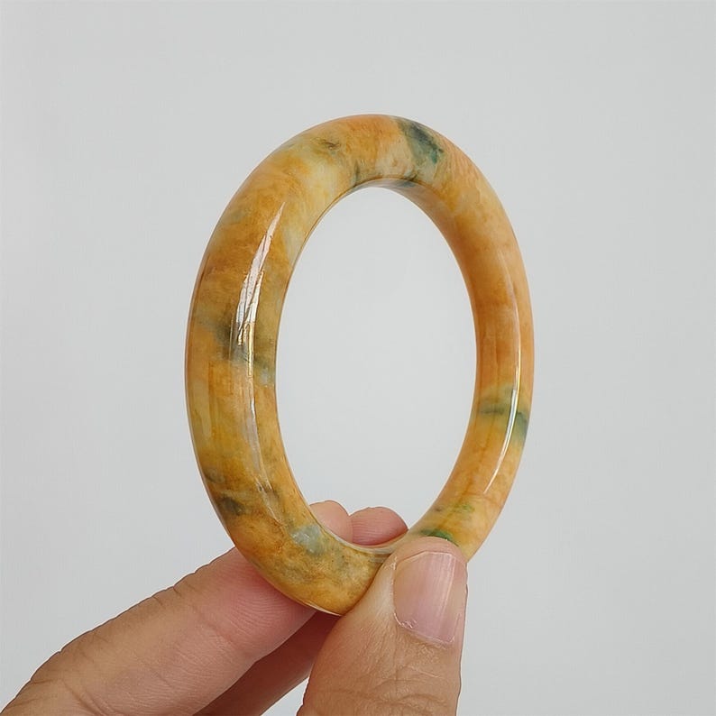 May include: A polished jade bangle bracelet. The bracelet is a warm yellow-orange color with green and white veining. The smooth, rounded surface reflects light, highlighting the natural variations in the stone.