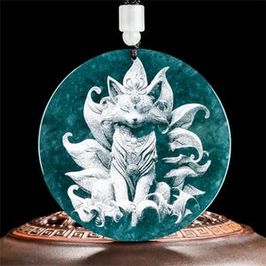May include: A teal-colored pendant with a detailed carving of a fox with multiple tails. The fox is centered on a circular jade stone, with a black cord and white bead for hanging. The pendant is displayed on a decorative stand.