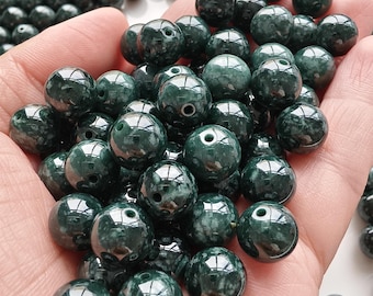 May include: A collection of round, polished dark green beads with mottled white and grey inclusions. Each bead features a central drilled hole, showcasing a smooth, glossy finish and a spherical shape, suitable for jewellery making and crafting projects.