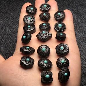 May include: A collection of 16 black wooden beads with intricate carvings and turquoise accents. Each bead features a unique design, including floral motifs, geometric patterns, and abstract shapes.