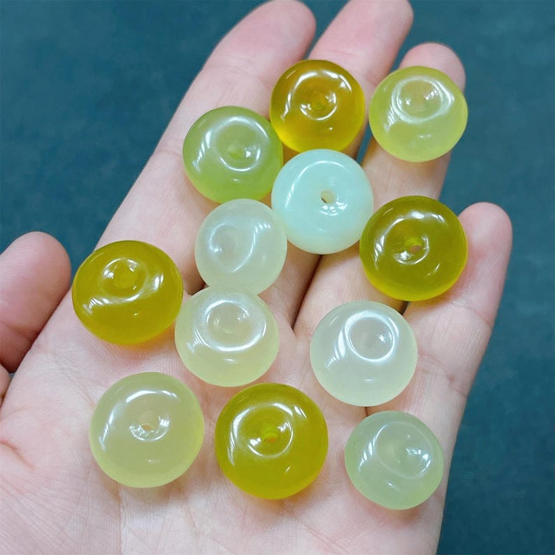 May include: A collection of 12 round, donut-shaped jade beads in shades of green and yellow. The beads have a hole in the center and are arranged on a hand.