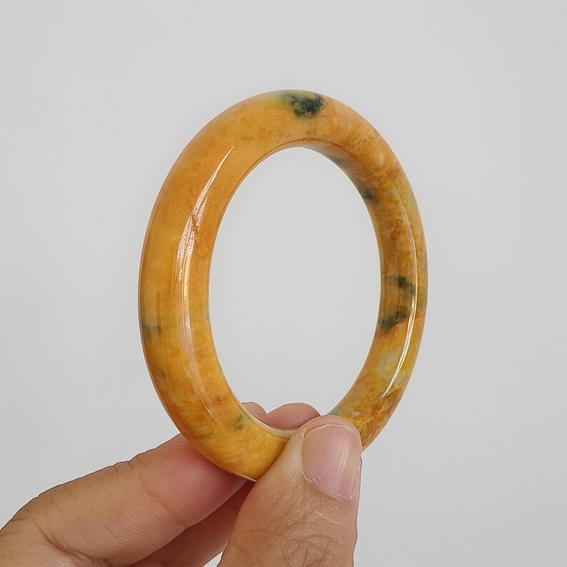 May include: A polished, oval-shaped jade bangle bracelet. The bracelet is a warm, golden-yellow color with subtle green and brown veining. The smooth surface reflects light, highlighting the natural variations in the stone.
