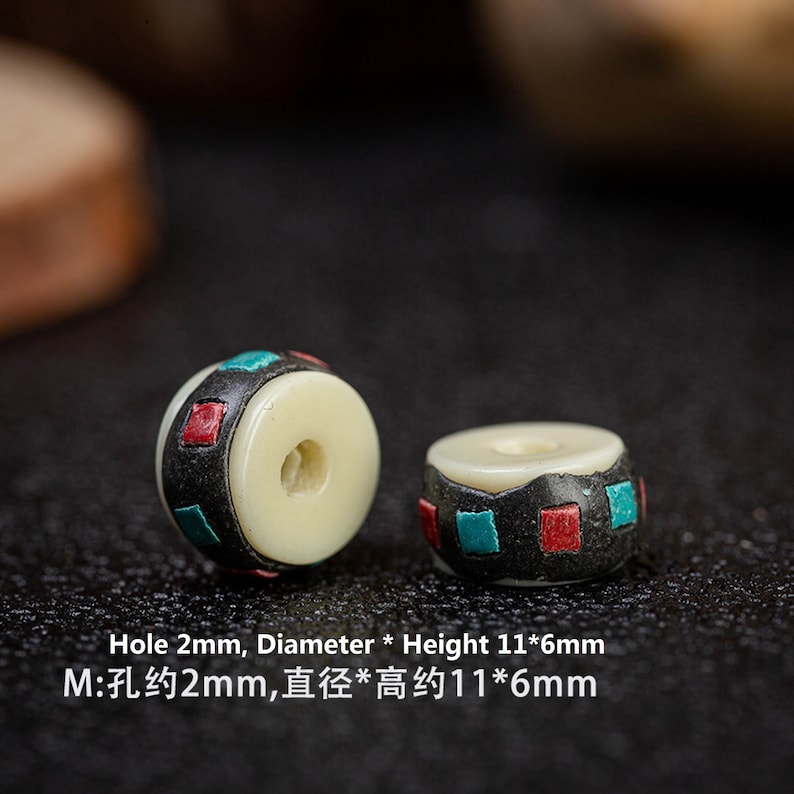 May include: Two white bone beads with black, red, and blue geometric patterns. The beads have a hole in the center and measure approximately 11 mm in diameter and 6 mm in height. The hole is approximately 2 mm in diameter.