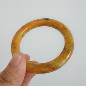 May include: A polished, round bangle bracelet in shades of yellow and brown. The bracelet has a smooth, glossy finish and appears to be made of stone. The bangle is held by a hand against a white background. Jewelry.