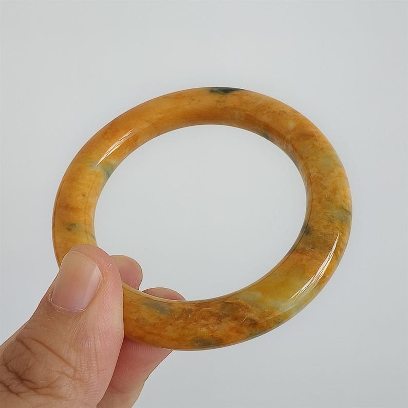 May include: A polished, round bangle bracelet in shades of yellow and brown. The bracelet has a smooth, glossy finish and appears to be made of stone. The bangle is held by a hand against a white background. Jewelry.