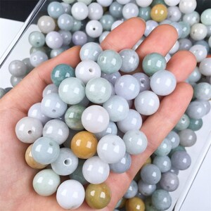 May include: A collection of round, smooth, jade green beads in various shades of green, white, and yellow. The beads are arranged in a hand, showcasing their size and texture.
