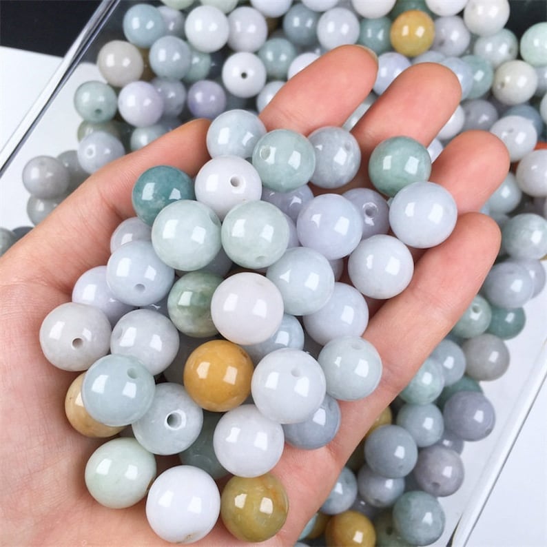 May include: A collection of round, smooth, jade green beads in various shades of green, white, and yellow. The beads are arranged in a hand, showcasing their size and texture.