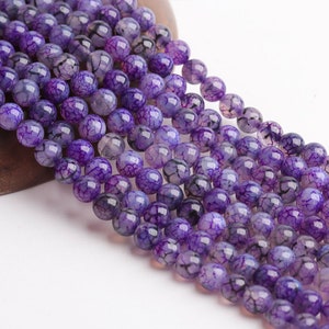 May include: Close-up of strands of round, purple gemstone beads. The beads have a cracked, web-like pattern in a deep violet colour with lighter purple and black veining. Ideal for jewellery making, each bead is approximately 0.8 cm in diameter.