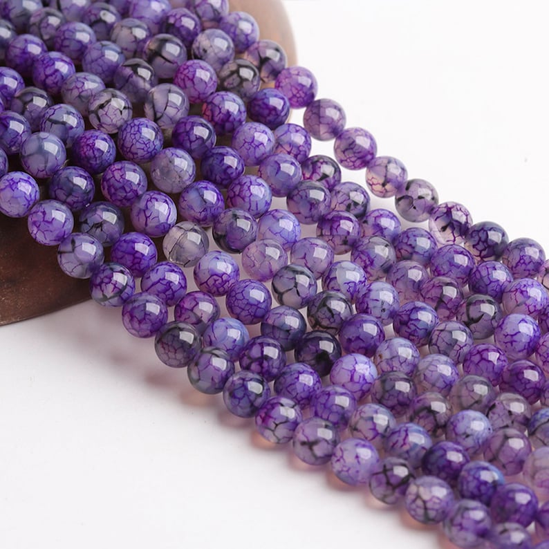 May include: Close-up of strands of round, purple gemstone beads. The beads have a cracked, web-like pattern in a deep violet colour with lighter purple and black veining. Ideal for jewellery making, each bead is approximately 0.8 cm in diameter.