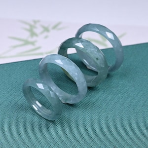 May include: Four jadeite rings stacked on top of each other. The rings are a pale green color and have a faceted design. The rings are on a green fabric surface.