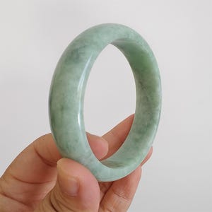 May include: A pale green jade bangle. The polished surface of the bracelet shows a subtle variation in colour, with areas of lighter and darker green. The circular shape of the bracelet is clearly visible.