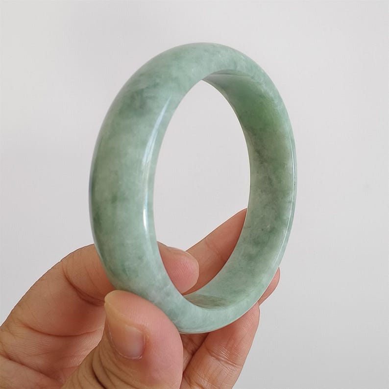 May include: A pale green jade bangle. The polished surface of the bracelet shows a subtle variation in colour, with areas of lighter and darker green. The circular shape of the bracelet is clearly visible.