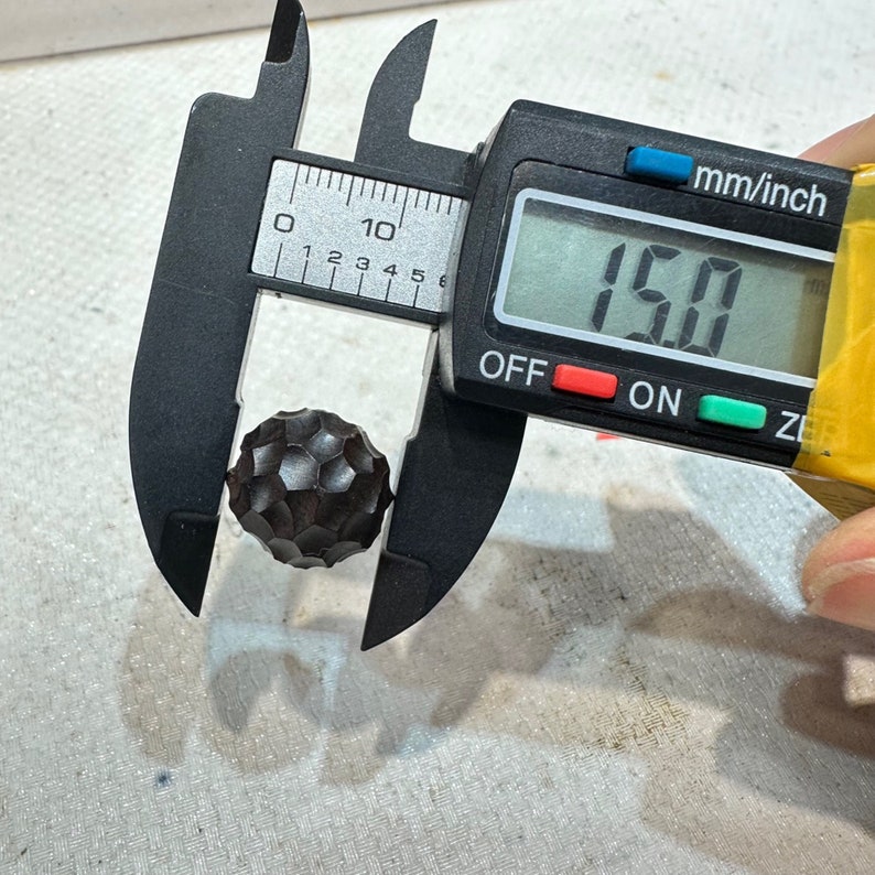 May include: A black metal ball with a textured surface is being measured with a digital caliper. The caliper reads 15.0 mm.