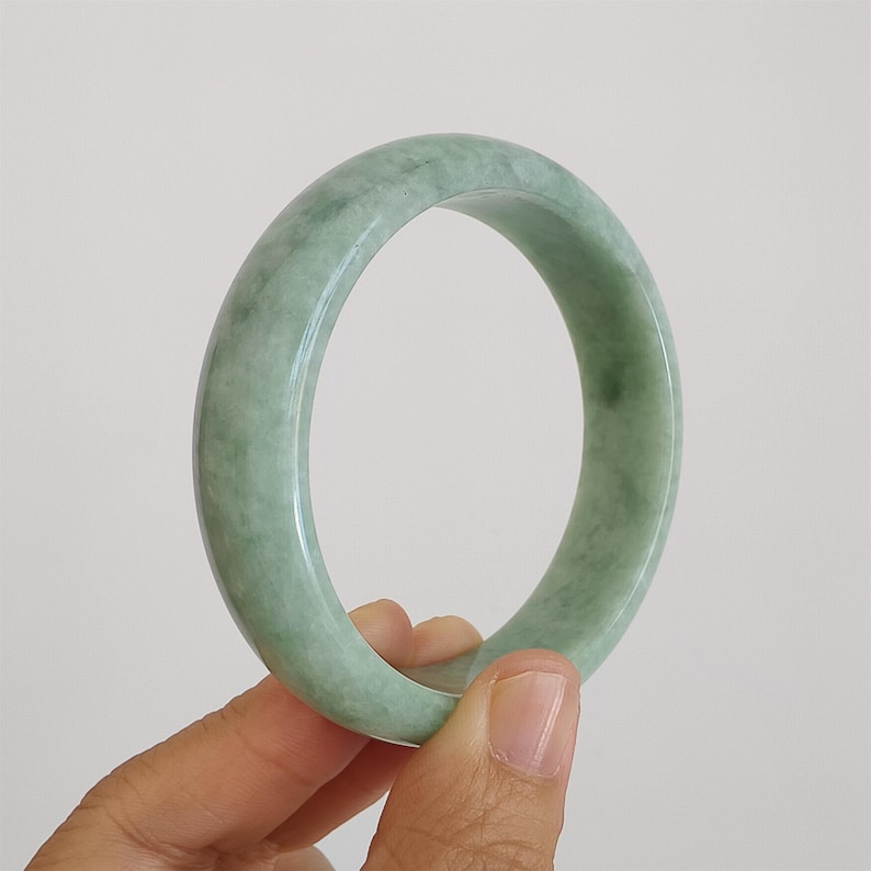 May include: A light green jade bangle. The polished surface of the bracelet shows variations in colour, with areas of lighter and darker green. The circular bracelet is held by a hand against a plain background.