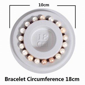 May include: A white, gray, and brown pearl bracelet on a white display stand. The bracelet measures 18 cm in circumference. The stand is 10 cm in diameter.